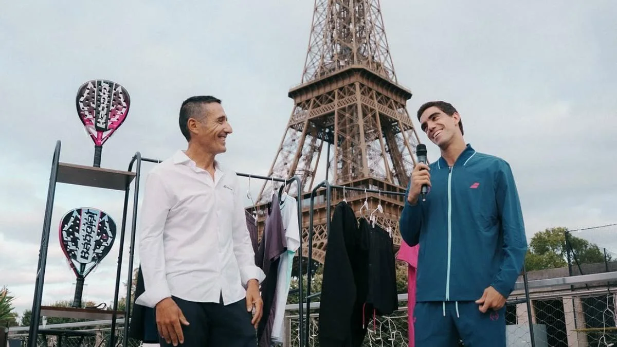 Juan Lebrón con un micrófono junto a un responsable de Babolat en la presentación, con la Torre Eiffel al fondo, un expositor con dos palas oscuras y rosas y una percha con la ropa de la colección