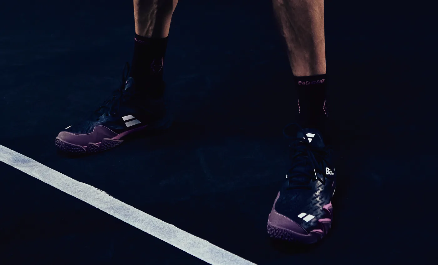 Detalle de los pies de Juan Lebrón sobre la pista: zapatillas Babolat azul marino con suela morada, calcetines negros con el logotipo en rosa y pantalón morado